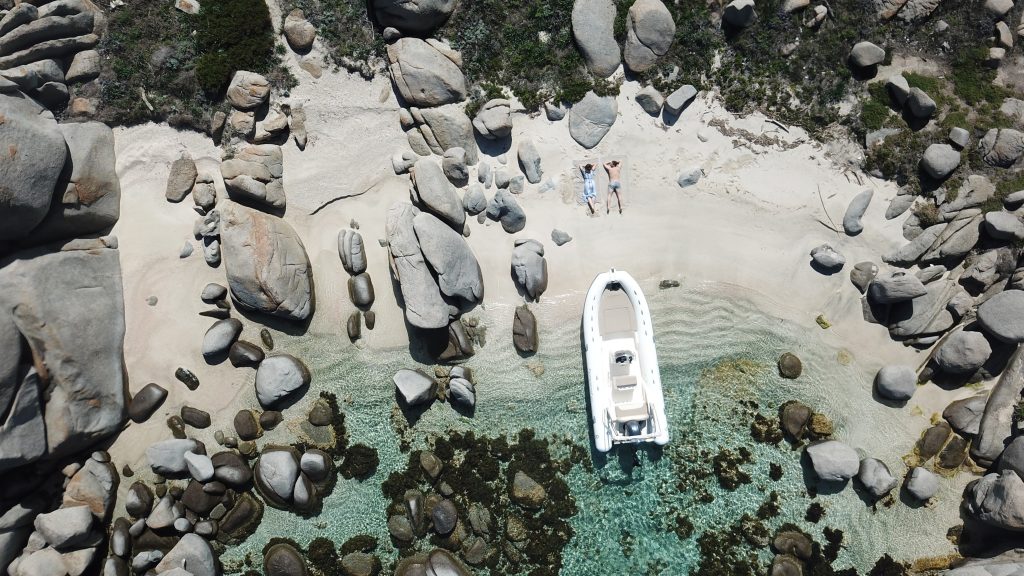 Vue aérienne d'un semi rigide, déposé sur le sable d'une crique sauvage par un couple, allongé sur le sable en arrière plan