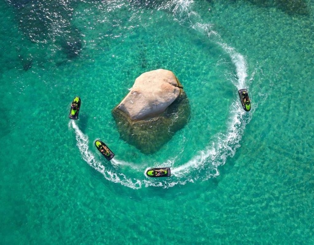 Vue aérienne de 4 jet skis noir et vert contournant un rocher dans une eau de mer translucide