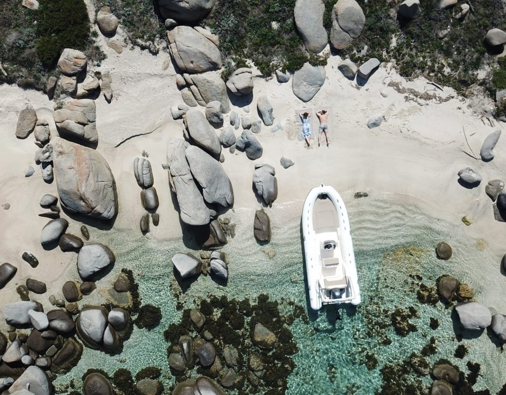 Vue aérienne d'un semi rigide, déposé sur le sable d'une crique sauvage par un couple, allongé sur le sable en arrière plan