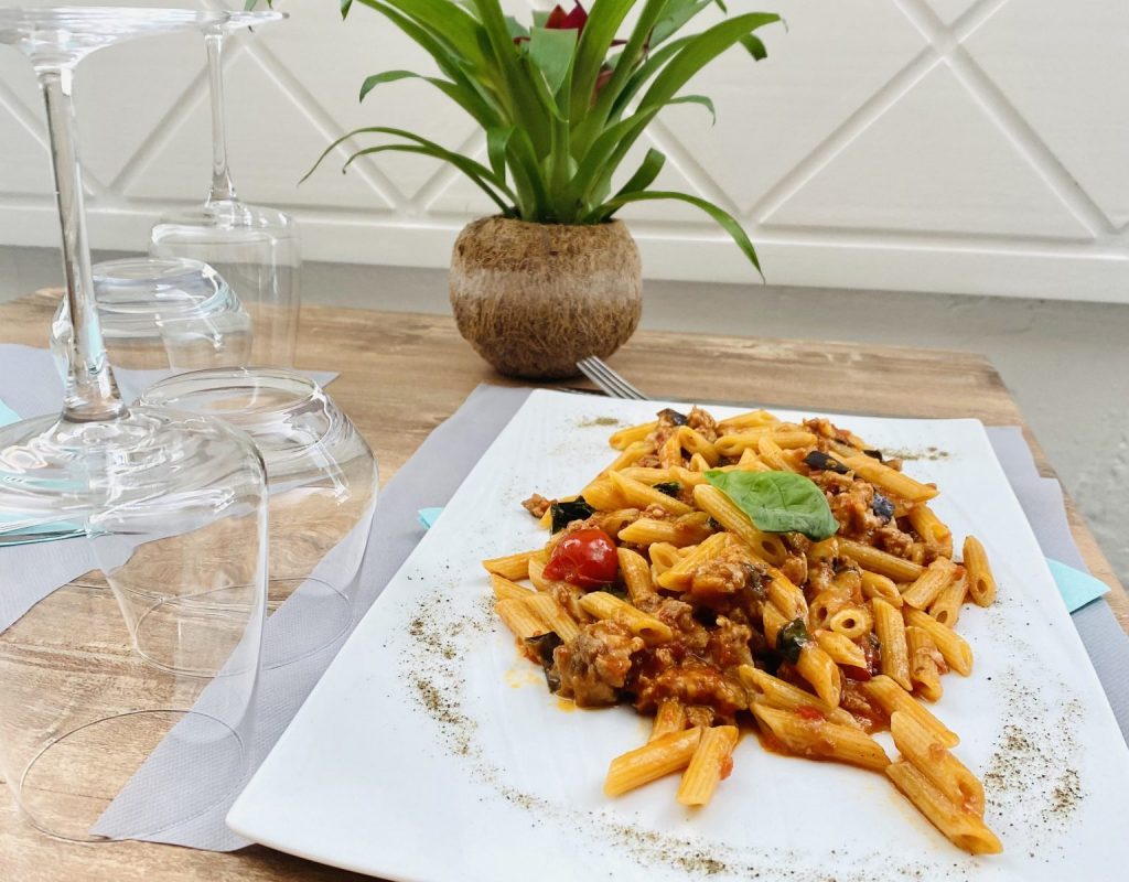 Table dressée à l'intérieur du restaurant avec assiette de penne et viande en sauce, tomates cerises et feuille de basilic