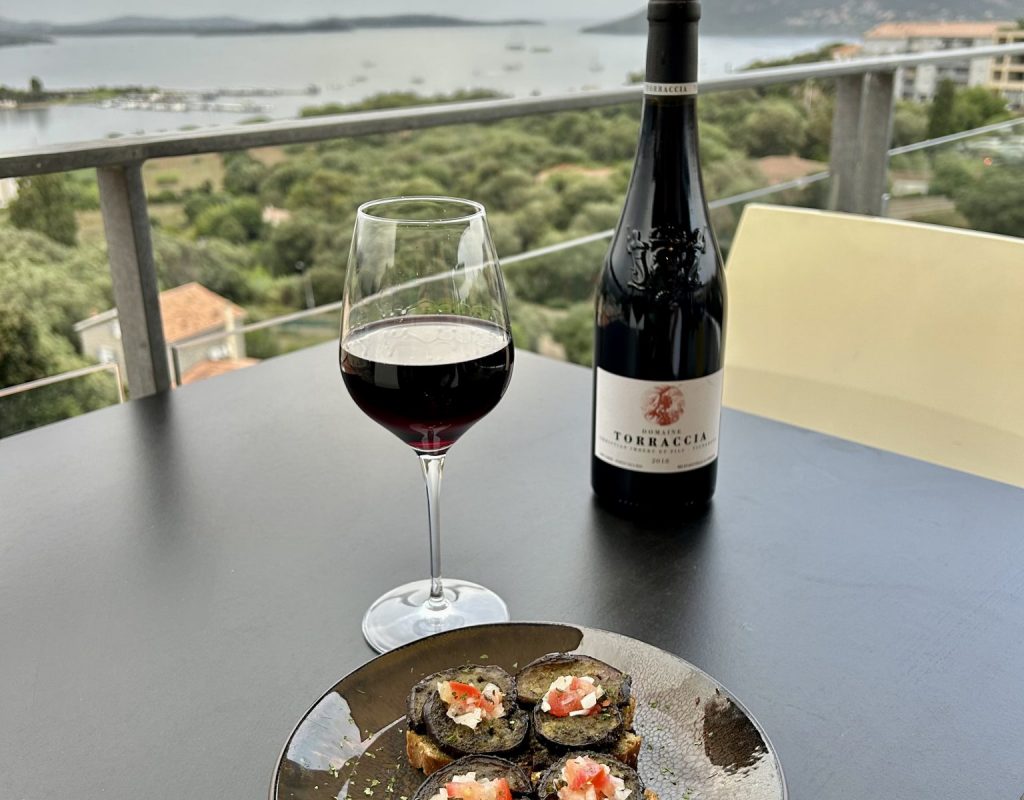 Table avec assiette de tapas, verre et bouteille de vin rouge, sur la terrasse extérieure, avec vue sur le golfe de Porto Vecchio