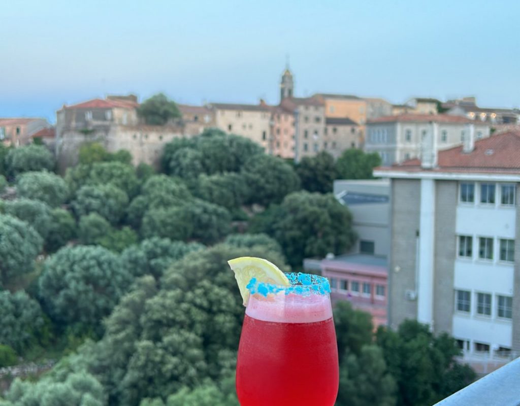 Cocktail de couleur rouge avec rondelle de citron décorative, posé sur le rebord de la terrasse extérieure, avec la ville de Porto Vecchio en arrière plan