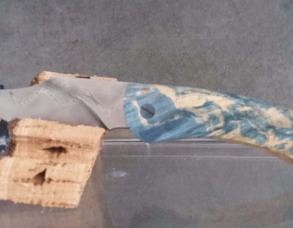 Zoom sur un couteau dont la lame est posée sur un morceau de bois, avec un manche en bois bleu et naturel
