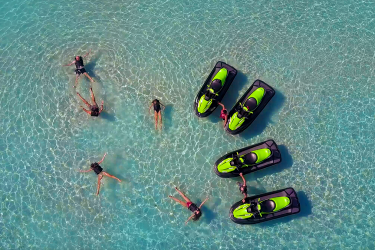 Vue aérienne avec 5 personnes allongées dans la mer cristalline à gauche, et à droite, 2 personnes, chacune entourée de 2 jet skis noir et vert