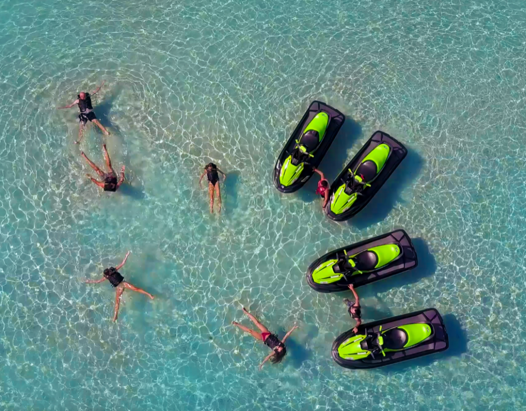 Vue aérienne avec 5 personnes allongées dans la mer cristalline à gauche, et à droite, 2 personnes, chacune entourée de 2 jet skis noir et vert