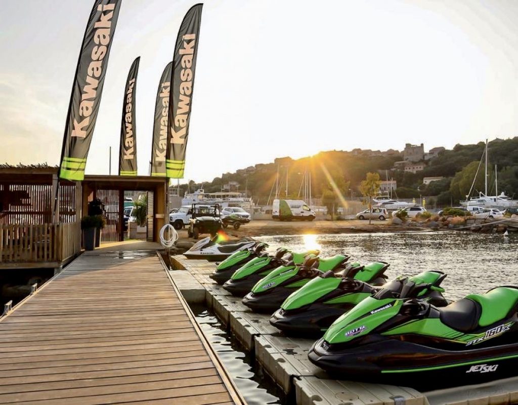 A gauche, ponton en bois menant au bureau d'accueil de Botti Racing sur le Port de Plaisance de Porto Vecchio, et sur la droite, 6 jet skis et le parking et la vue sur la haute ville en arrière plan