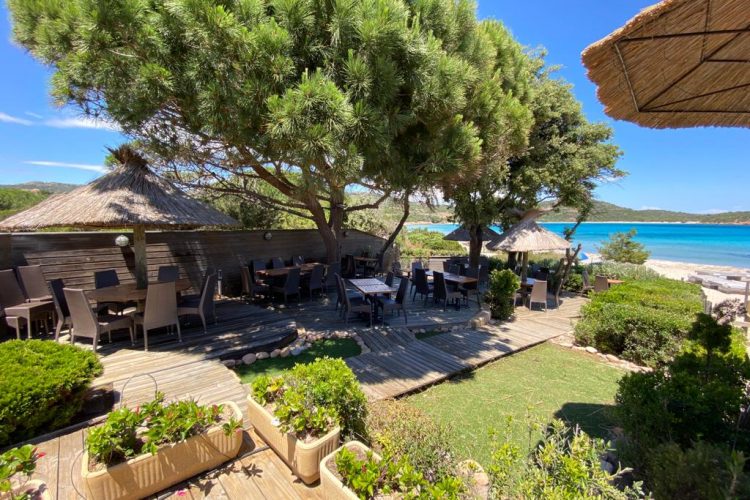 Tables dressées pour le service sur la terrasse du restaurant, en bois et ombragée, avec vue sur la baie de Rondinara, par une journée ensoleillée