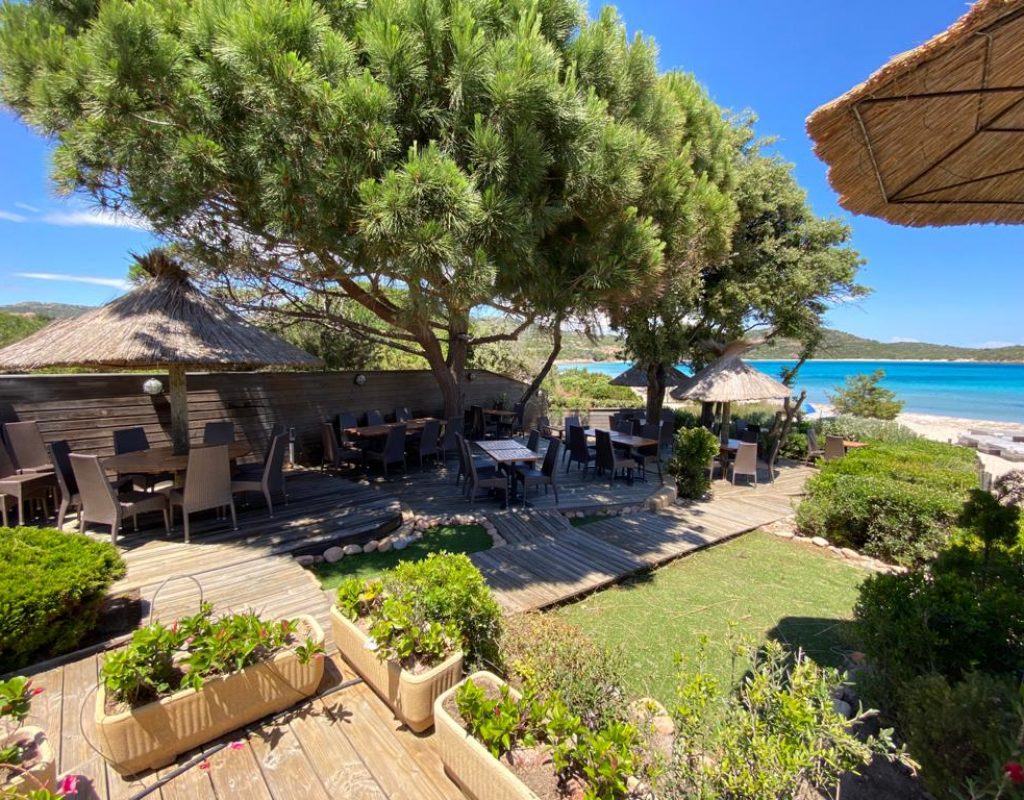 Tables dressées pour le service sur la terrasse du restaurant, en bois et ombragée, avec vue sur la baie de Rondinara, par une journée ensoleillée