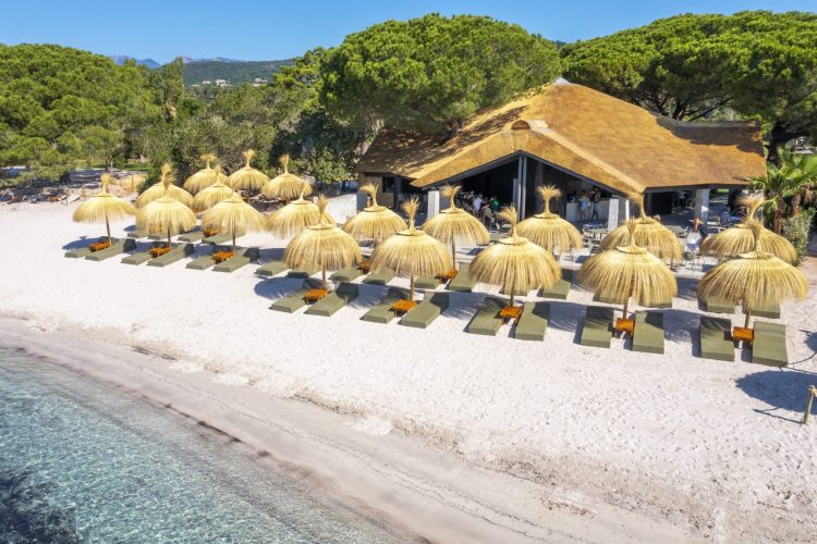 Vue aérienne, de côté, de la paillotte ensoleillée avec son restaurant et ses transats et parasols au bord de l'eau