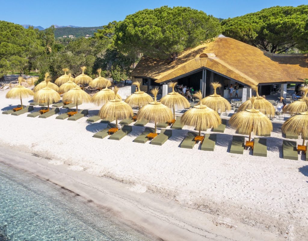 Vue aérienne, de côté, de la paillotte ensoleillée avec son restaurant et ses transats et parasols au bord de l'eau