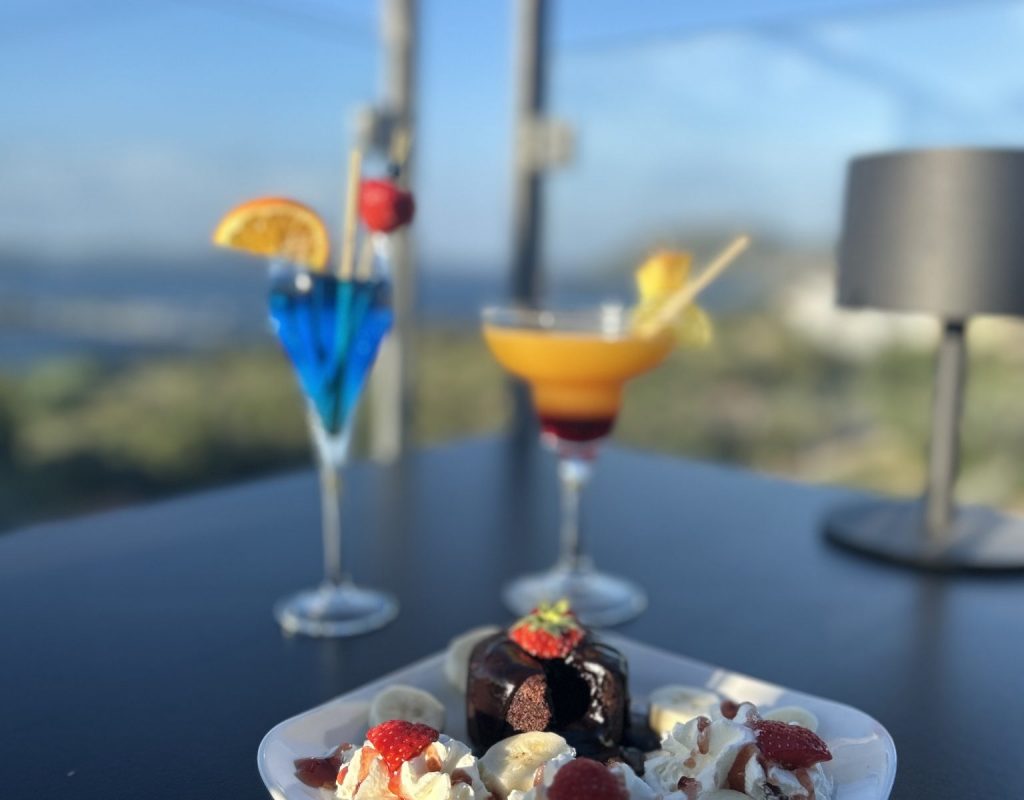 Assiette sucrée avec gateau au chocolat, morceaux de bananes et de fraises, chantilly, avec cocktails et golfe de Porto Vecchio en arrière plan
