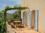 The stone patio with a grapevine roof is in the famous Corsican town Pigna. There are a wooden table with two benches and a mountain peak in the background.